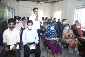 A briefing session in Bangladesh: a man speaking into a microphone to a seated room of young candidates holding forms.