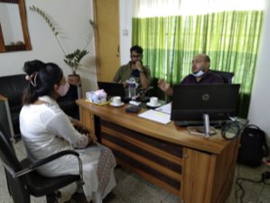 Three people in an office meeting around a desk with a laptop, discussing a hiring decision.