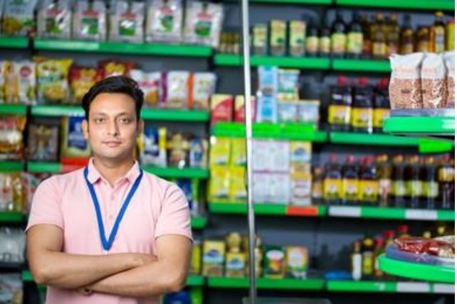 A young retail staff member in a pink polo shirt and lanyard standing in front of stocked grocery shelves.