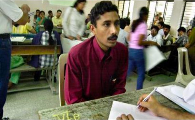 A young man at a desk in a crowded assessment hall in Bangladesh, speaking with an assessor reviewing papers.