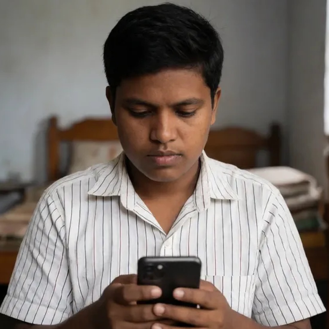 A young neurodivergent Bangladeshi learner wearing headphones, focused on a mobile phone.